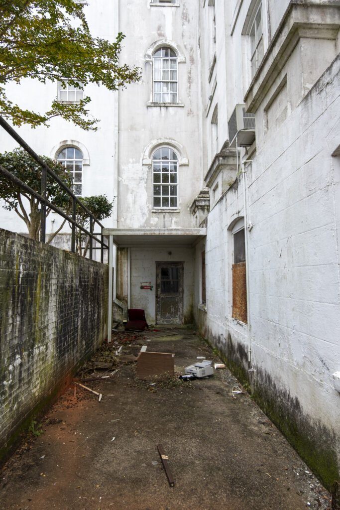 Decaying hallway outside the Powell Building at Central State Hospital in Milledgeville, Georgia, with abandoned chairs scattered on the floor and peeling walls showing years of neglect.